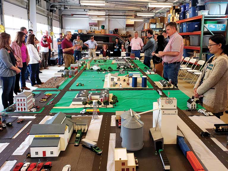 Photograph of about a dozen people in a multi-bay garage viewing a long scale model of an agricultural town with a grain elevator in the foreground and farmland in the background