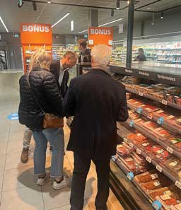 Three people in professional attire looking at packaged fresh pork cuts in an open multi-level refrigerated display case