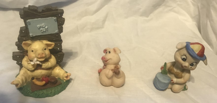 Three pig figurines - one eating a banana with the words "Pig Out" on a sign, one in a bikini with a pail and shovel, one on its back in a cute pose.